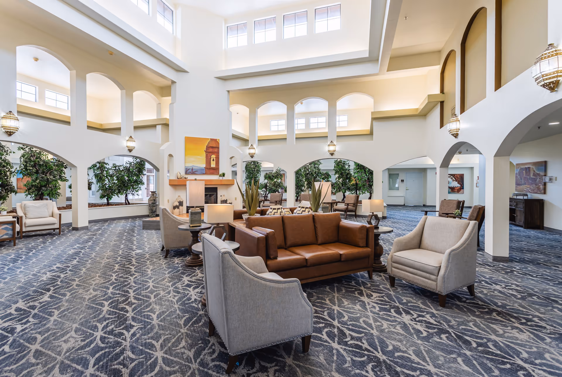 Spacious and well-lit senior living facility common area with high ceilings, arched openings, and large windows. The room features a brown leather sofa, beige armchairs, side tables with lamps, potted plants, and decorative artwork on the walls. The carpet has a blue and white pattern.