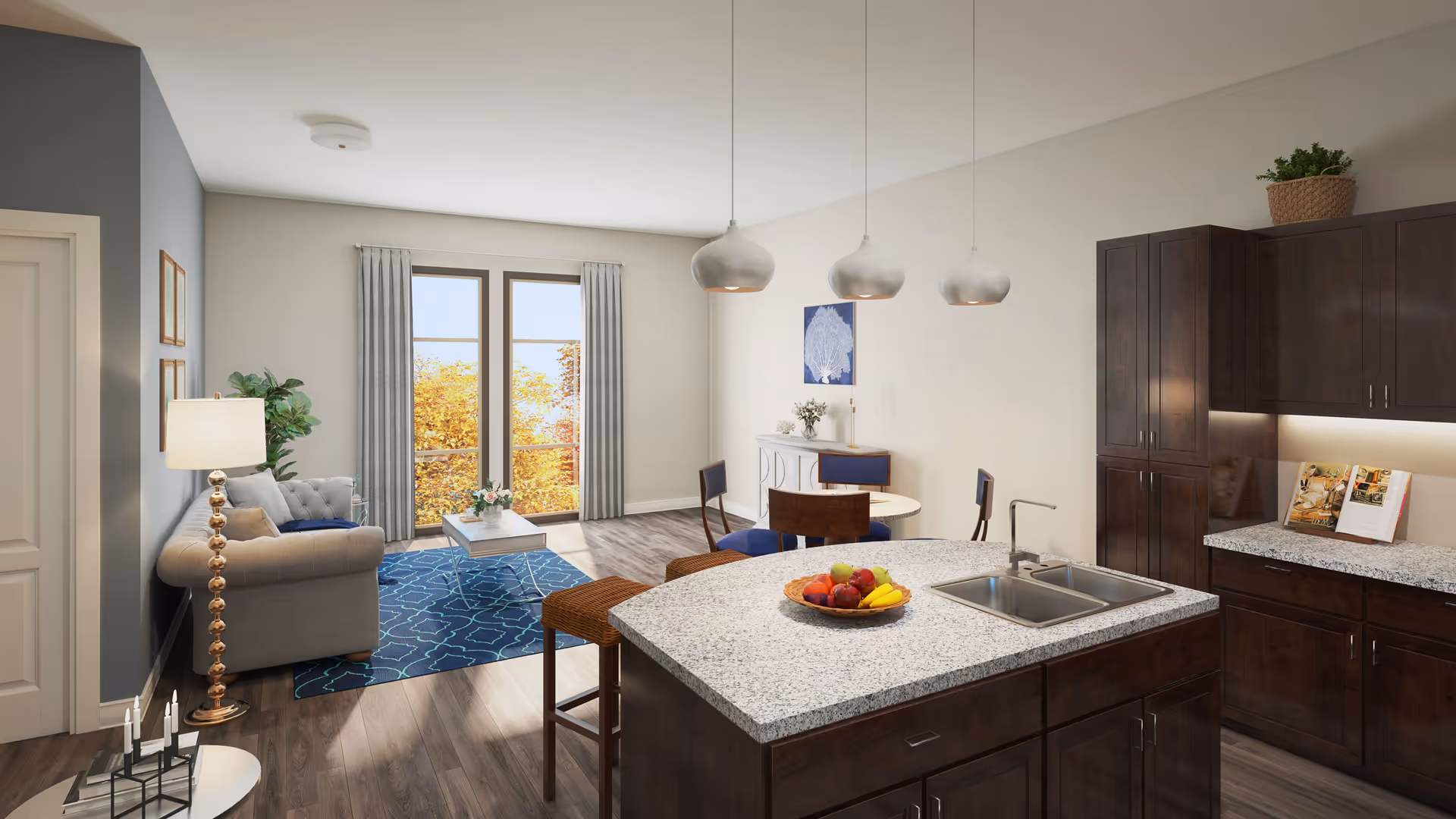 A modern open-concept living space featuring a kitchen island with a granite countertop and a bowl of fruit, dark wood cabinets, a dining area with a round table and four chairs, and a living room with a beige tufted sofa, a blue patterned rug, a coffee table, and large windows with gray curtains letting in natural light.