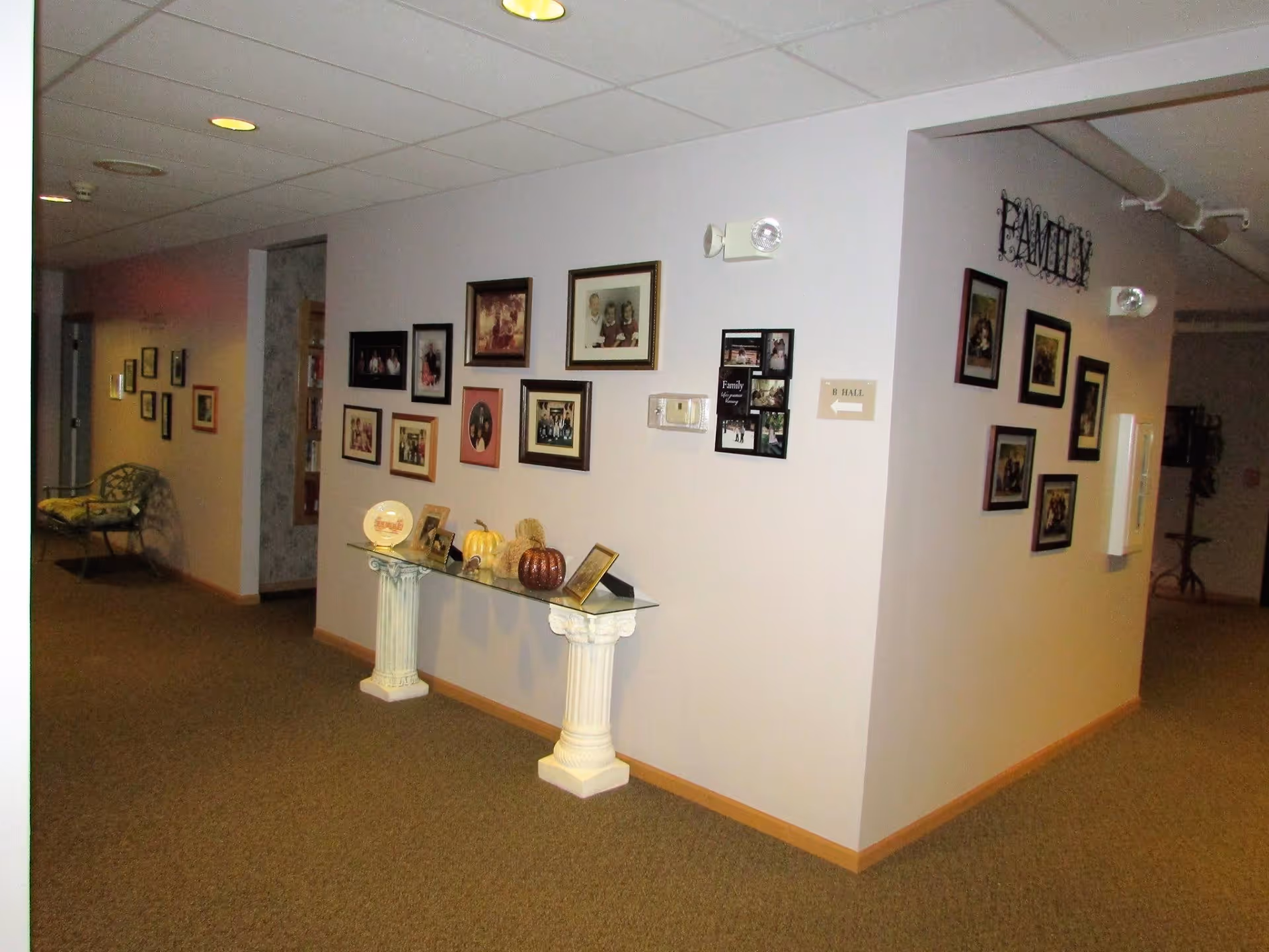 Interior hallway of Eastland Retirement Community with beige carpet and light-colored walls decorated with multiple framed family photos. A glass table supported by two white decorative columns holds small decorative items including pumpkins. A bench with a cushion is visible down the hallway.