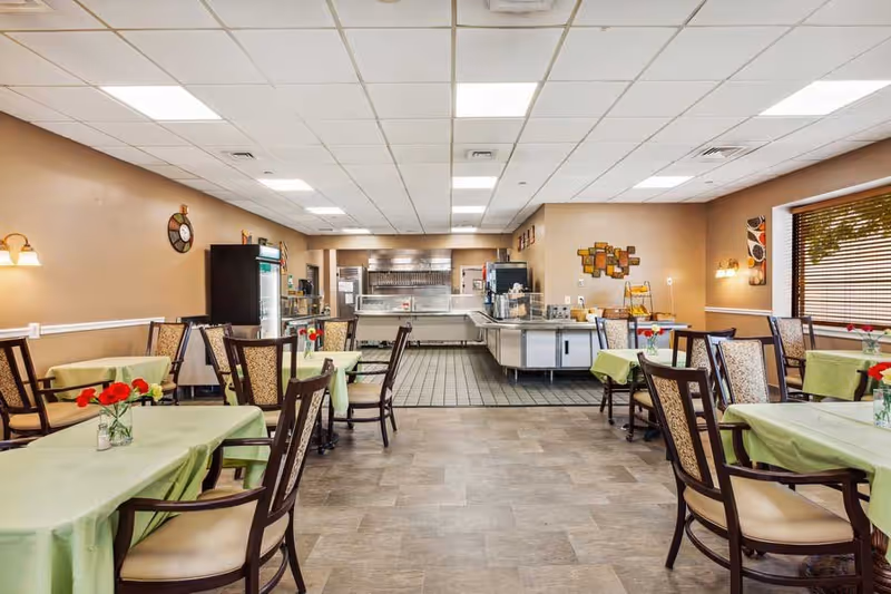 A dining room in a senior living facility with several tables covered in light green tablecloths and decorated with small vases of red flowers. The room has beige walls, a tiled floor near the serving area, and wooden chairs with cushioned seats. The serving area at the back includes a stainless steel counter and kitchen equipment. Large windows with blinds allow natural light into the room.