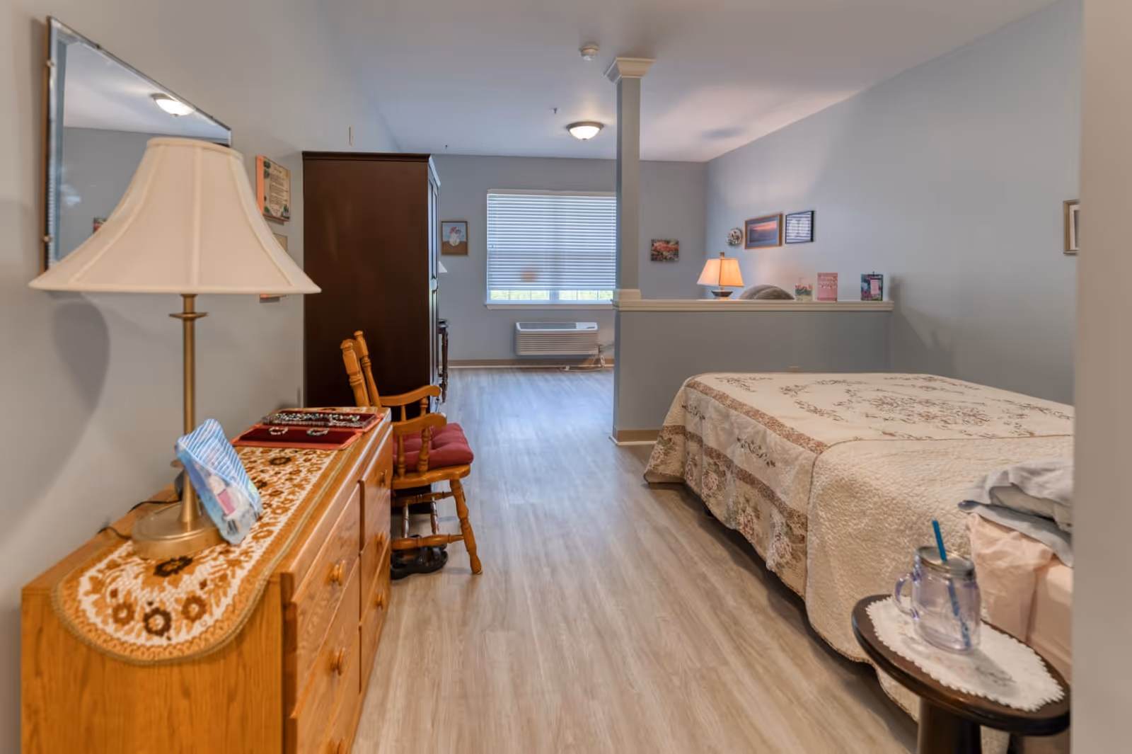 A senior living facility bedroom with a bed covered in a floral quilt, a wooden dresser with a lamp and decorative cloth, a wooden chair with a red cushion, and a small round table holding a glass with a straw. The room has light-colored wood flooring, light blue walls, and a window with blinds letting in natural light. There is a half wall separating the bed area from a small sitting area with a lamp and framed pictures on the wall.
