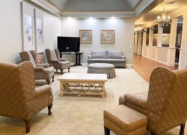 A cozy living room area in Shawnee Memory Care featuring several upholstered armchairs and a sofa arranged around a wooden coffee table and a round ottoman. A flat-screen TV is placed on a small table against the wall, which is decorated with framed floral artwork. The room has beige carpeting, soft lighting, and an open hallway with white wainscoting and wooden flooring leading to other areas.