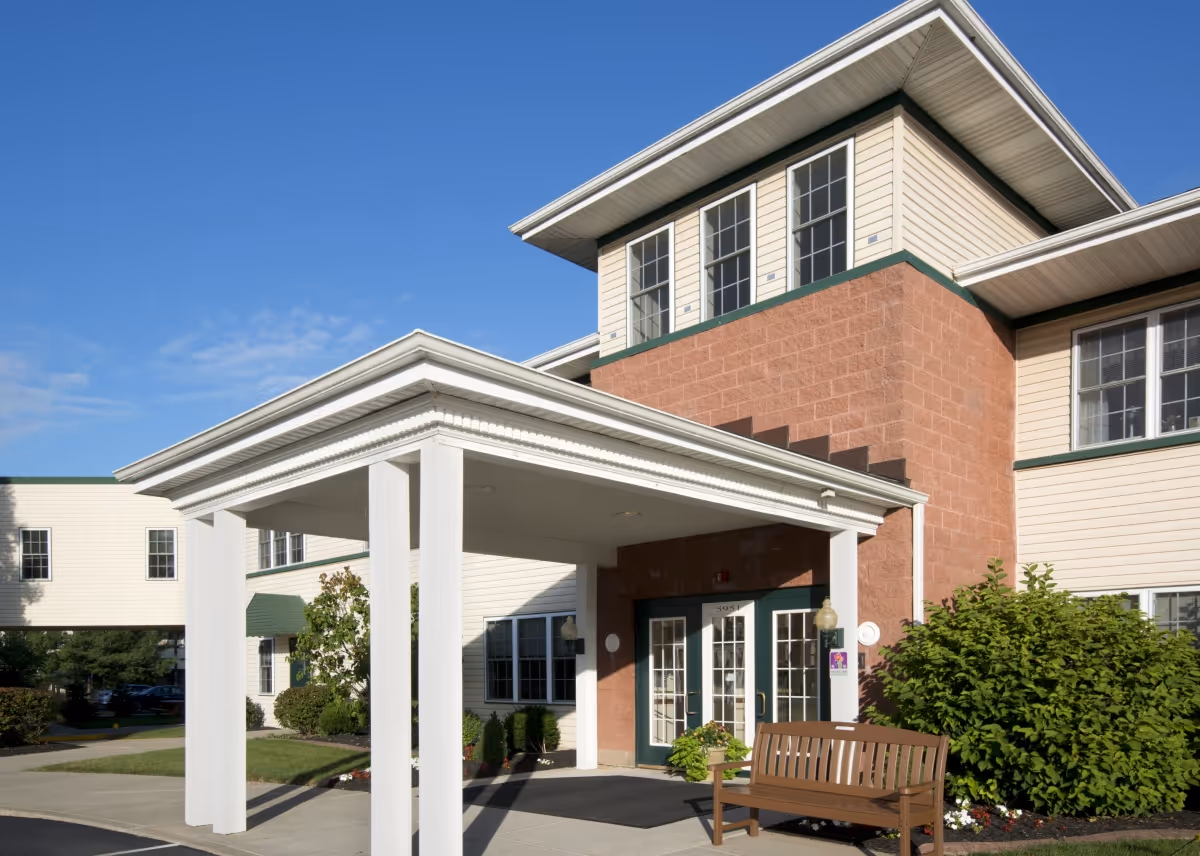 Front entrance of a multi-story senior living building with a covered portico, bench, and landscaping under a clear blue sky.