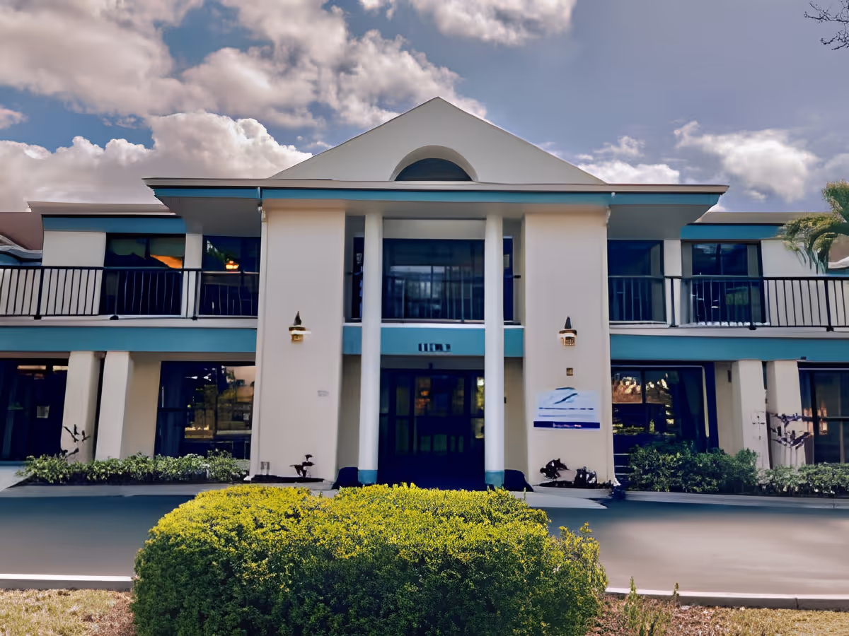 Front exterior of a two-story cream and teal senior living building with columns, balconies, and shrub landscaping under a partly cloudy sky.