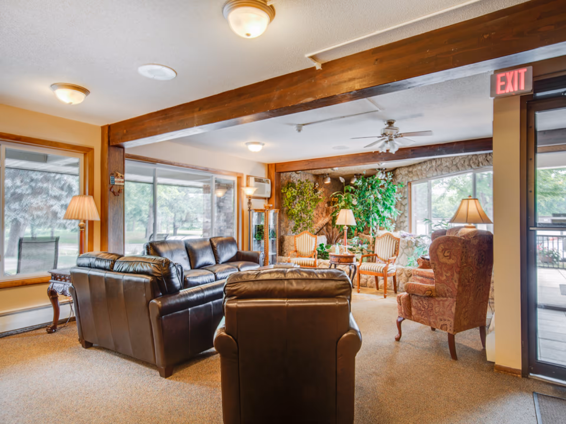 A cozy living room area with leather sofas and armchairs arranged around a small table. The room features large windows with views of greenery outside, wooden beams on the ceiling, and several lamps providing warm lighting. There are also potted plants and a stone wall in the background, creating a welcoming atmosphere.