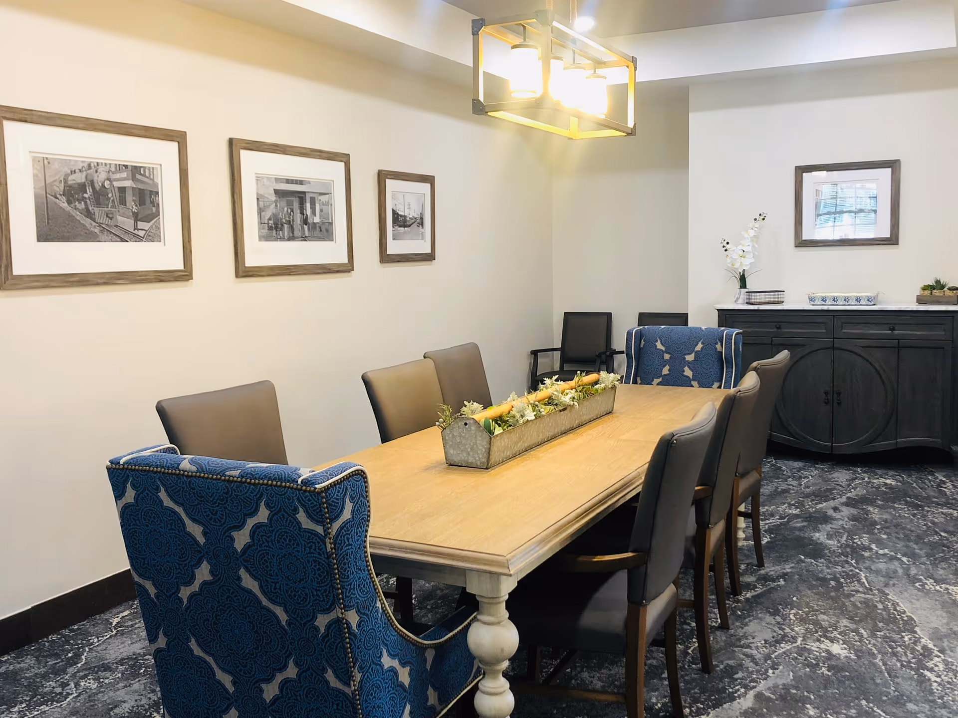 A dining room with a long wooden table surrounded by eight chairs, two of which have blue patterned upholstery. The room features a modern rectangular ceiling light fixture, framed black and white photographs on the walls, a dark wooden sideboard with decorative items, and a carpeted floor with a marbled pattern.