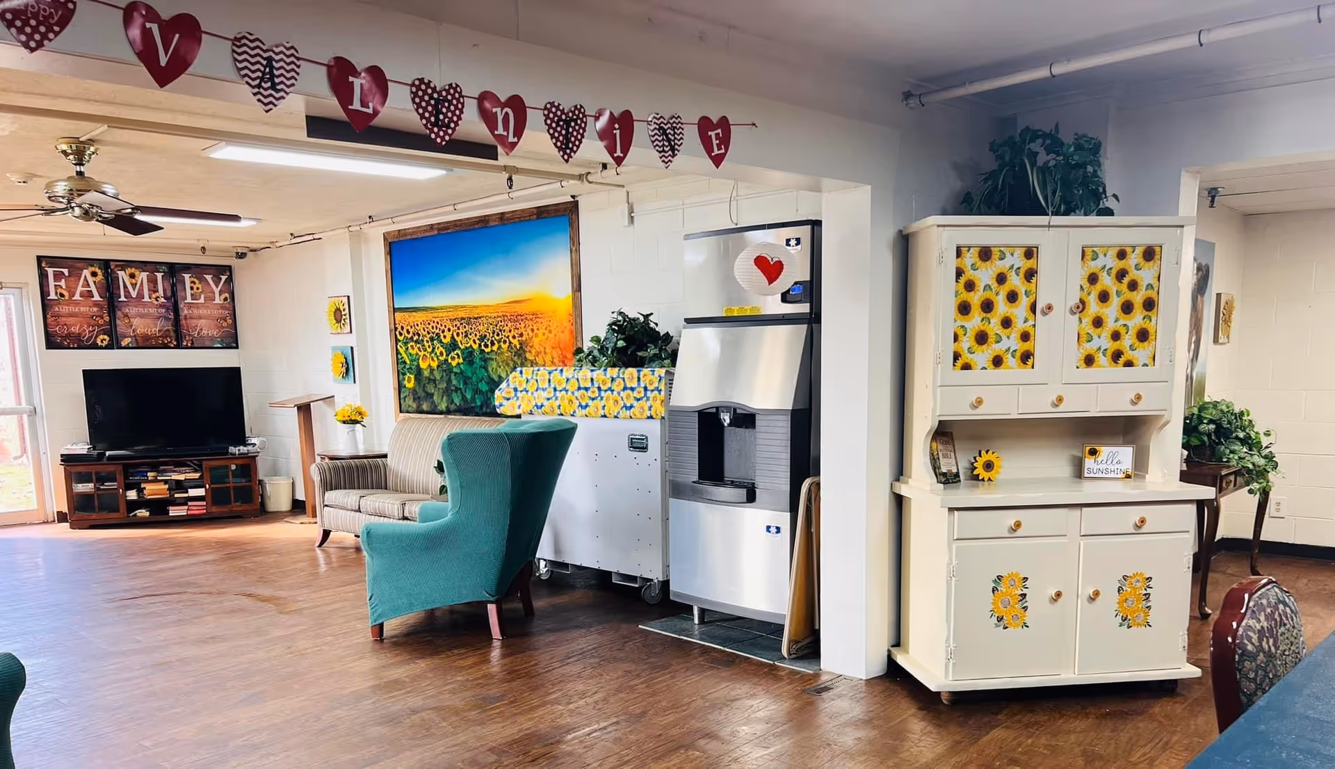 A cozy living room area in a nursing and rehab facility decorated with sunflower-themed artwork and furniture. There is a green armchair and a striped sofa near a large painting of a sunflower field at sunset. A wooden cabinet decorated with sunflower patterns stands next to a water dispenser. Above the room, a garland of red heart-shaped cutouts spells out 'Valentine'. A TV and media console are visible in the background, along with a wall decoration that reads 'FAMILY'.