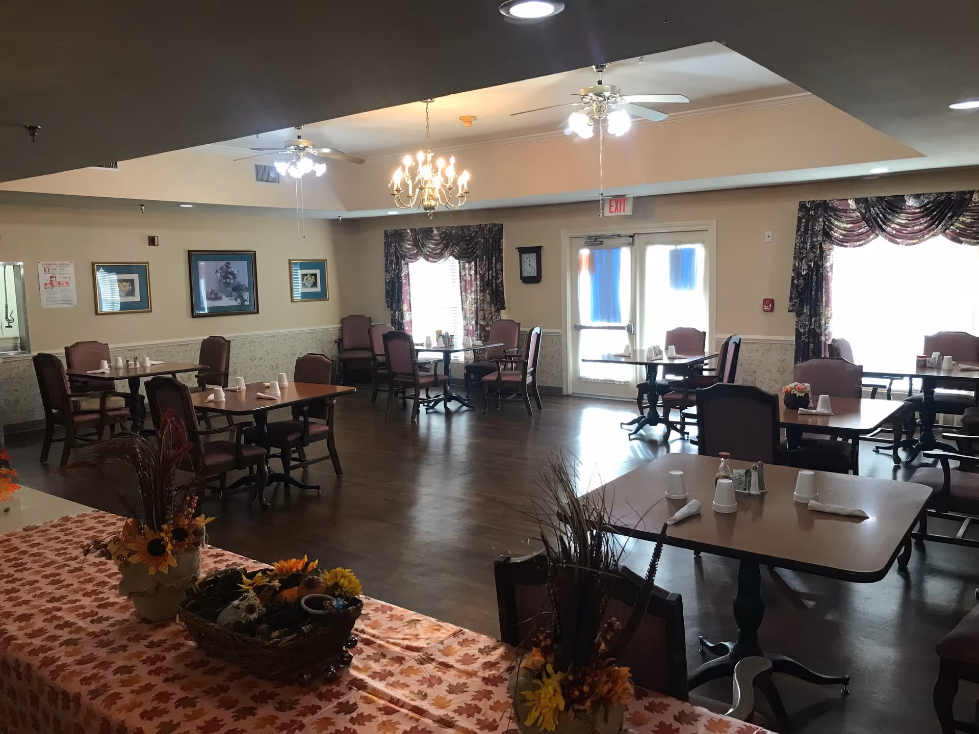 Dining room with multiple tables and chairs arranged neatly. Tables are set with white napkins and cups. The room has wooden flooring, floral curtains on the windows, framed pictures on the walls, and ceiling fans with lights. A chandelier hangs from the ceiling, and there are decorative flower arrangements on a table covered with a fall-themed tablecloth in the foreground.