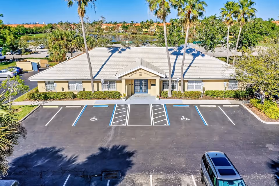 Front exterior view of a single-story building with a light-colored roof and beige walls, surrounded by palm trees and greenery. There is a parking lot in front with designated handicapped parking spaces and a few cars parked. The building entrance has double doors under a small peaked roof.