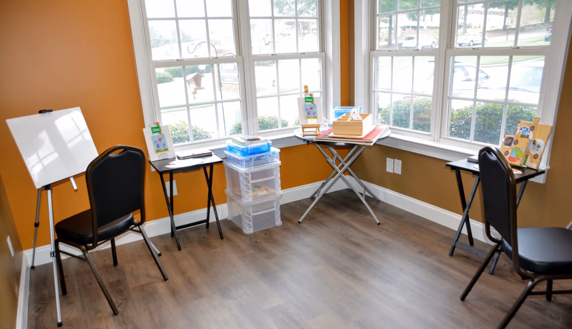 A small corner room with orange walls and large windows letting in natural light. The room contains two black chairs, two small black folding tables with craft supplies and books on them, a whiteboard on an easel, and clear plastic storage bins stacked in the corner.