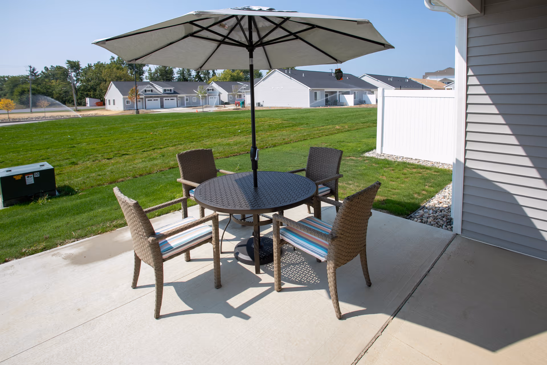Patio with a round table, umbrella and four wicker chairs on a concrete slab overlooking a grassy yard and neighboring single-story homes.
