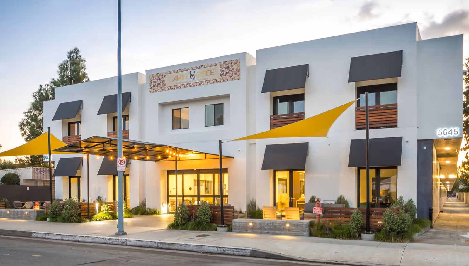 Exterior view of AvantGarde Senior Living facility showing a modern two-story building with white walls, black window awnings, yellow shade sails, and illuminated entrance at dusk.
