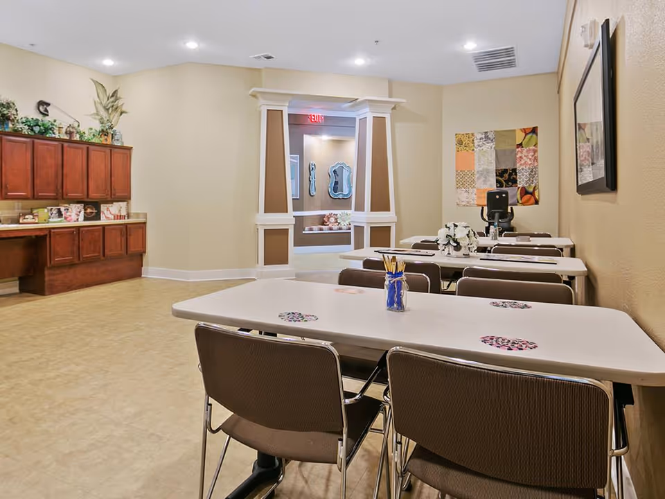 A well-lit room with several tables and chairs arranged in rows. The tables have small decorative items, including a jar with pencils and a flower arrangement. The walls are beige with framed artwork and a colorful patchwork quilt hanging. There is a wooden cabinet with plants and decorative items on the left side. In the background, there is an open doorway with white columns and a red exit sign above it.
