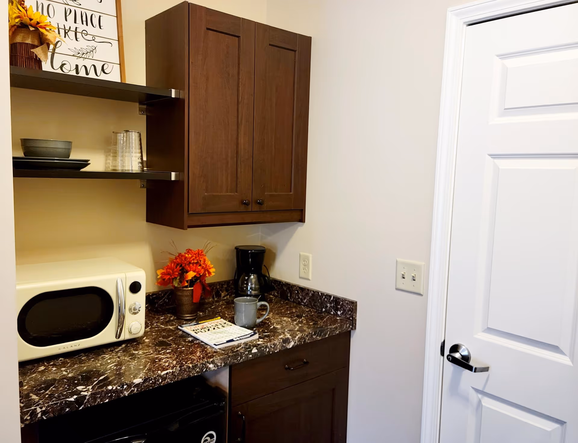 A small kitchenette area with a dark marble countertop, a white microwave, a coffee maker, a gray mug, a small potted plant with orange flowers, and a notepad. Above the countertop are two dark wooden cabinets and two shelves holding plates and glasses. A white door with a silver handle is visible on the right side.