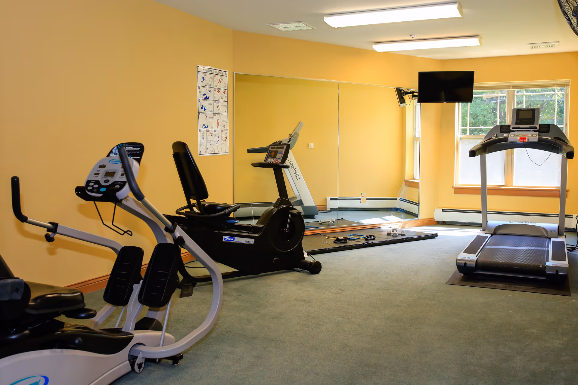 A small fitness room with exercise equipment including a treadmill, a recumbent bike, and an elliptical machine. The room has yellow walls, a large mirror on one side, a wall-mounted TV, and windows letting in natural light.