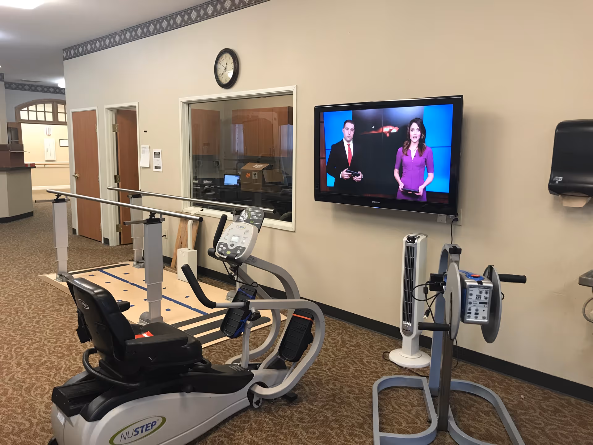 Rehabilitation room with a recumbent exercise bike, parallel bars, wall-mounted TV showing news anchors, and other therapy equipment.
