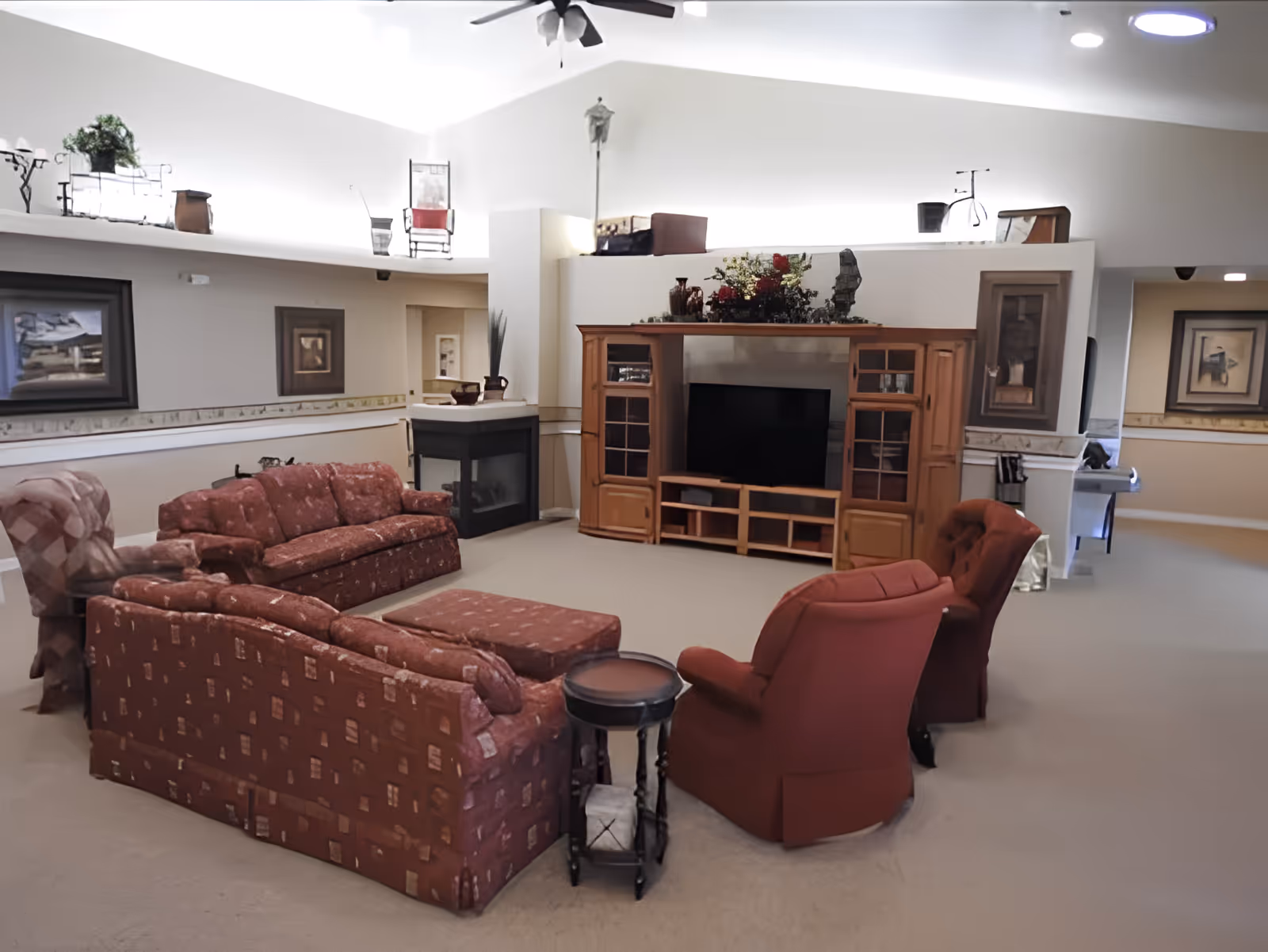A spacious living room with high vaulted ceilings, featuring a red patterned sofa set, two red armchairs, a wooden entertainment center with a TV, and a corner fireplace. The room is decorated with framed pictures on the walls and various decorative items on top of the entertainment center and ledges.