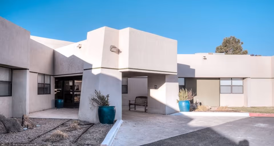 Exterior view of Bear Canyon Rehabilitation Center showing a single-story building with a covered entrance, large windows, two blue planters with plants, a bench under the covered area, and a clear blue sky.