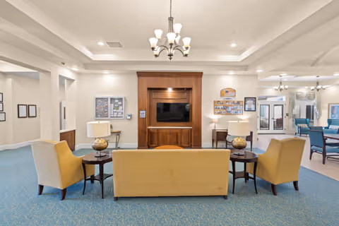 A spacious living room area in a memory care community featuring a yellow sofa and two matching armchairs arranged around a wooden entertainment center with a flat-screen TV. The room has blue carpet, beige walls, and ceiling lights including a chandelier. There are side tables with lamps on either side of the sofa, bulletin boards on the walls, and additional seating visible in the background.