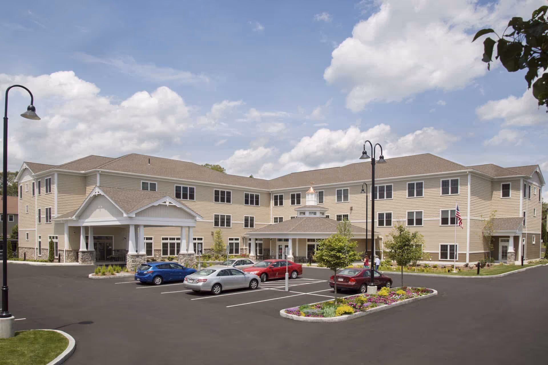 Exterior view of a three-story senior living facility building with beige siding and a covered entrance. There is a parking lot with several cars parked and a landscaped island with flowers and small trees. The sky is partly cloudy.