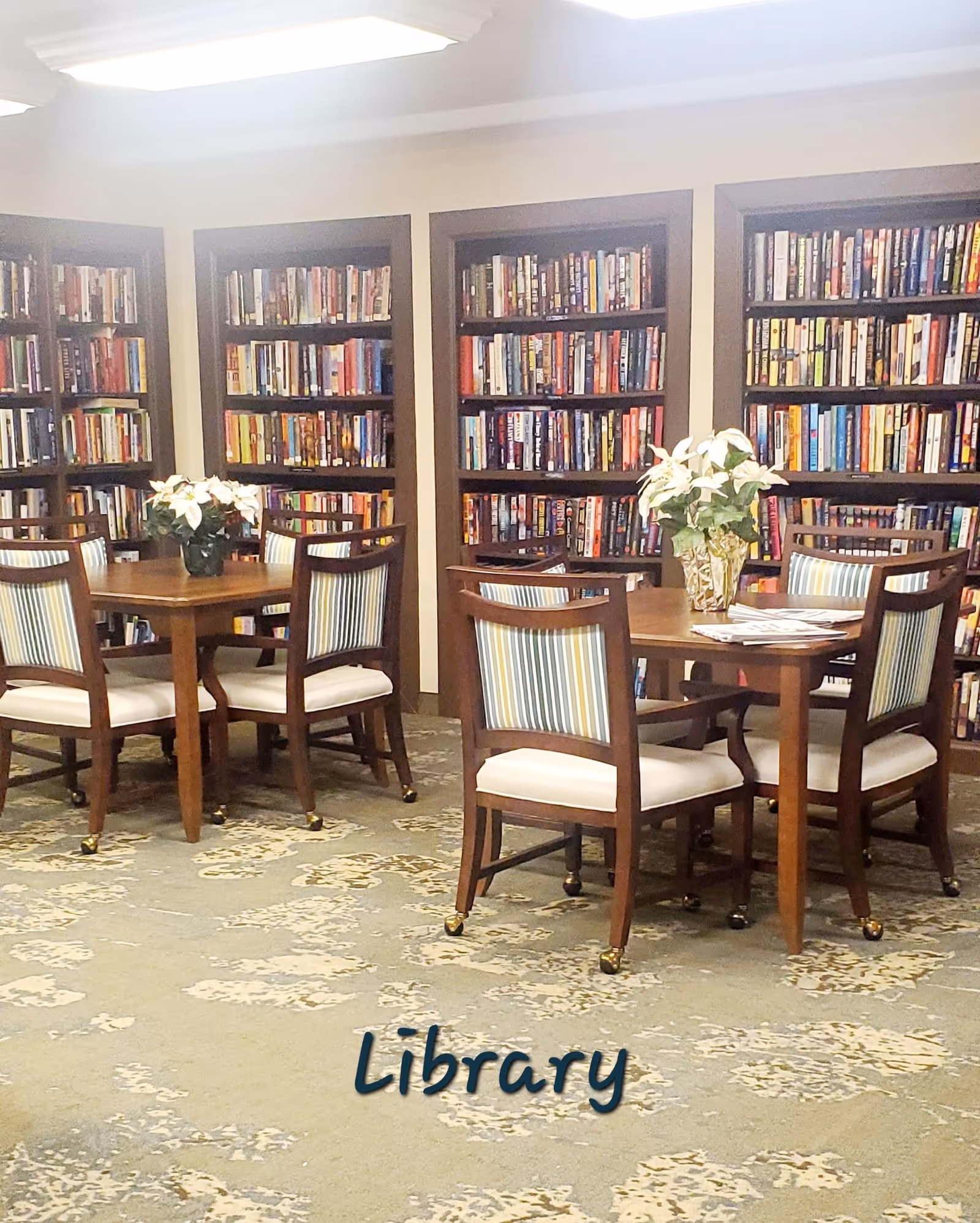 A library room with wooden bookshelves filled with books along the walls. Two wooden tables with four chairs each are placed on a patterned carpet. Each table has a vase with white flowers. The word 'Library' is written on the carpet in front of the tables.