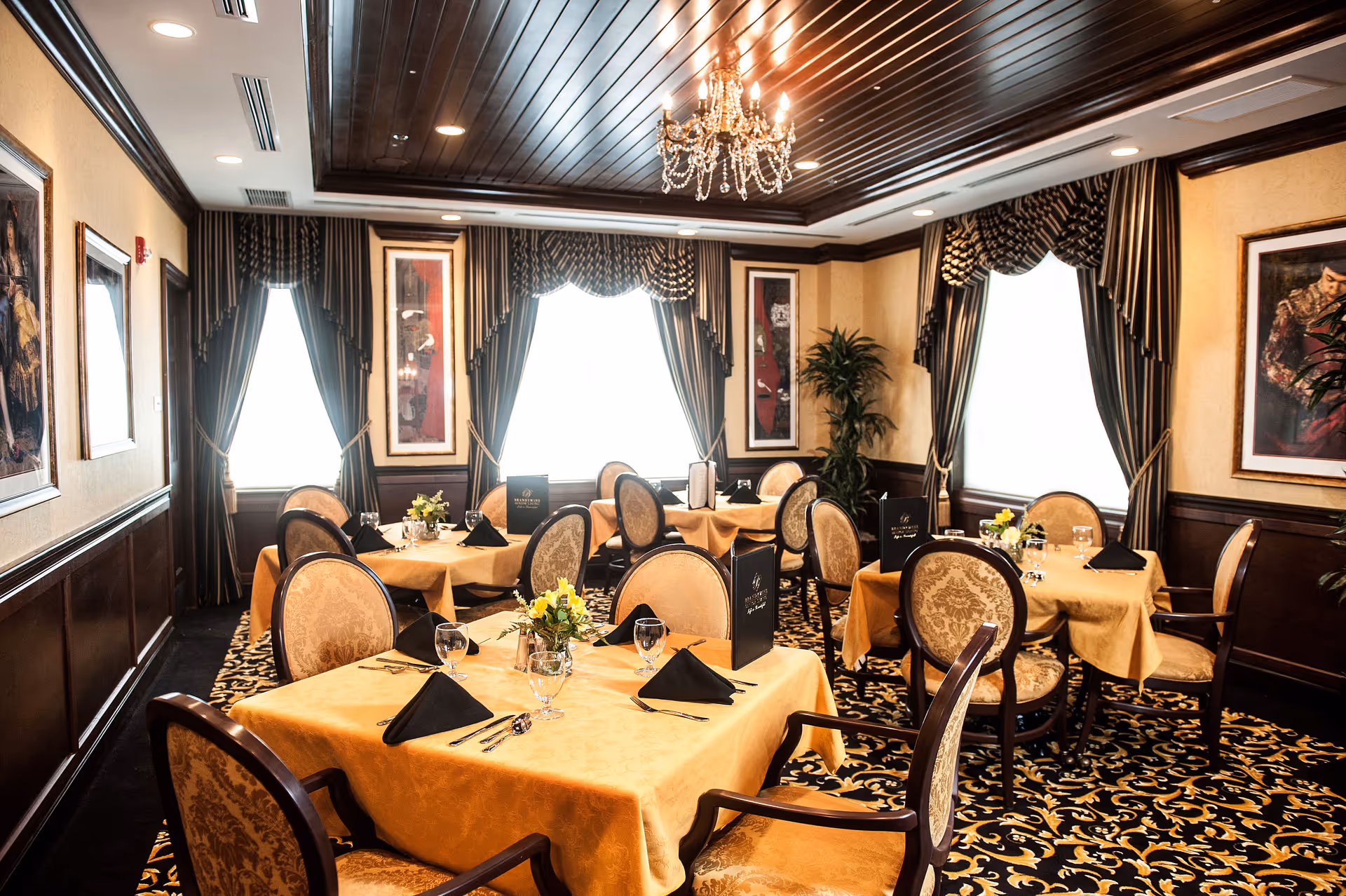 Elegant dining room with multiple tables covered in gold tablecloths, each set with black napkins, glassware, and silverware. The room features ornate chairs with patterned upholstery, large windows with dark drapes, framed artwork on the walls, a dark wood-paneled ceiling with recessed lighting, and a crystal chandelier hanging in the center.