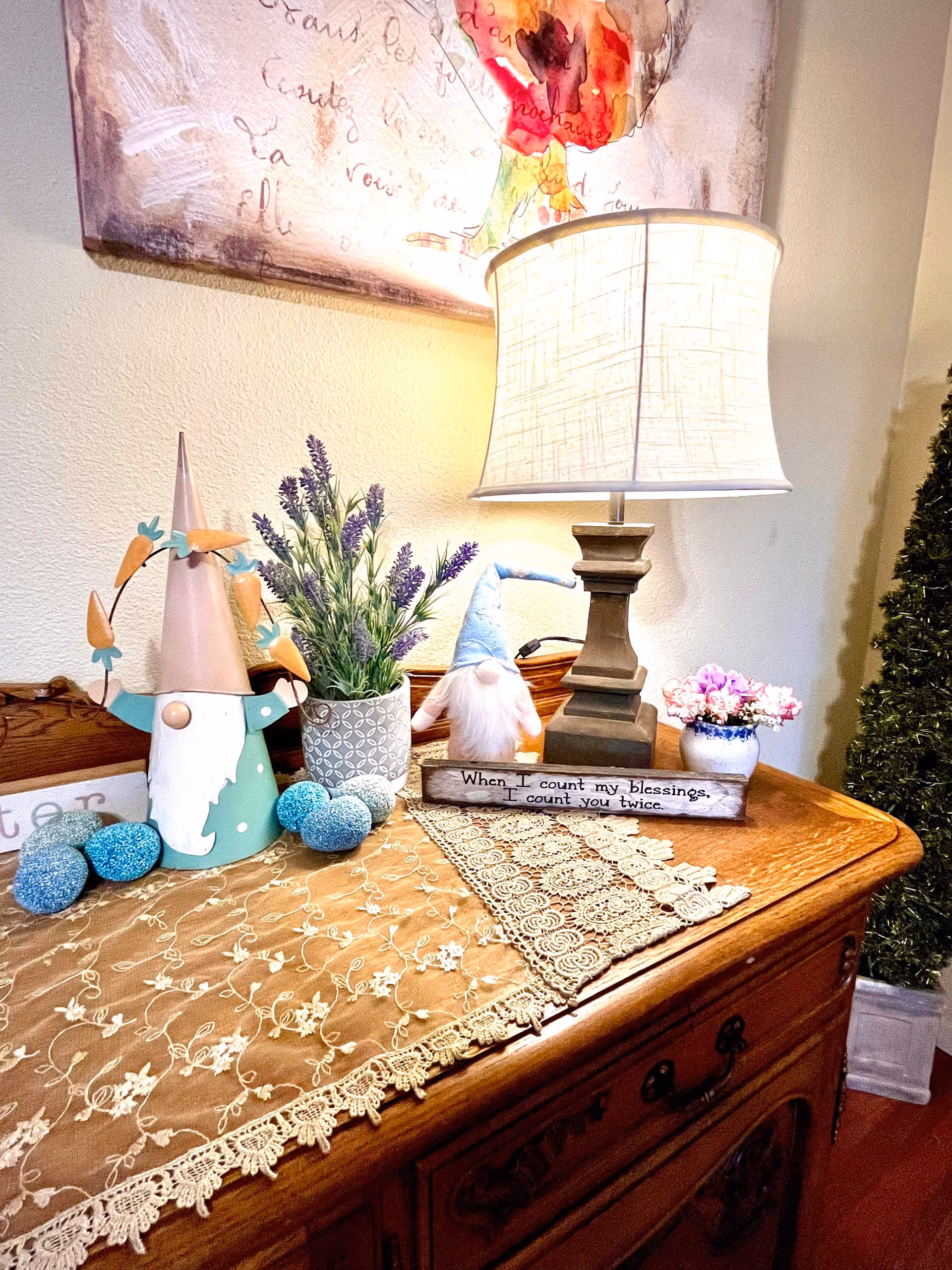 A wooden side table decorated with a beige lace table runner, a table lamp with a beige shade, two small gnome figurines, a potted lavender plant, a small vase with pink flowers, and several decorative blue eggs. A framed artwork with floral and script elements hangs on the wall above the table. A small sign on the table reads, 'When I count my blessings, I count you twice.'