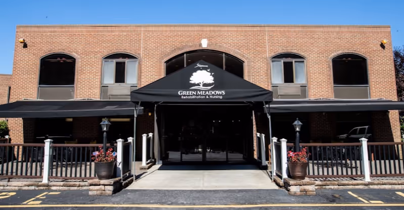 Front exterior view of a two-story brick building with a black awning displaying the Green Meadows Rehabilitation & Nursing logo above the entrance. There are windows on the upper floor and a small fenced patio area with potted plants on either side of the entrance.