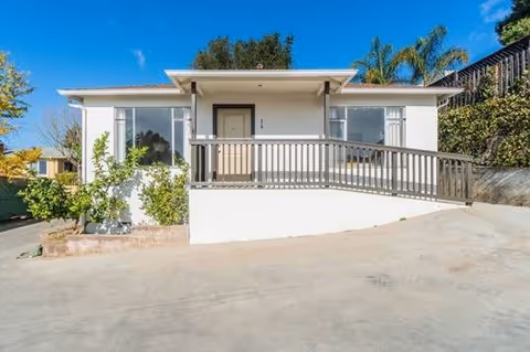 Small single-story white building with a front ramp and railing, concrete driveway, and palm trees under a blue sky.