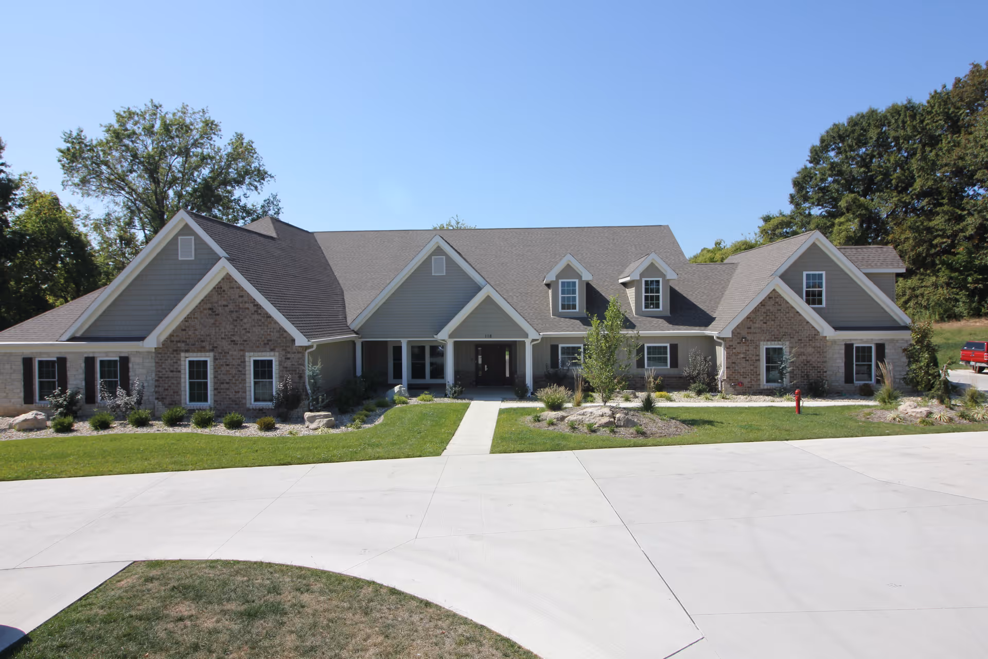 Front exterior view of a single-story residential building with a combination of brick and siding facade, multiple gabled roofs, and a concrete driveway leading to the entrance. The building is surrounded by green grass, small shrubs, and trees under a clear blue sky.