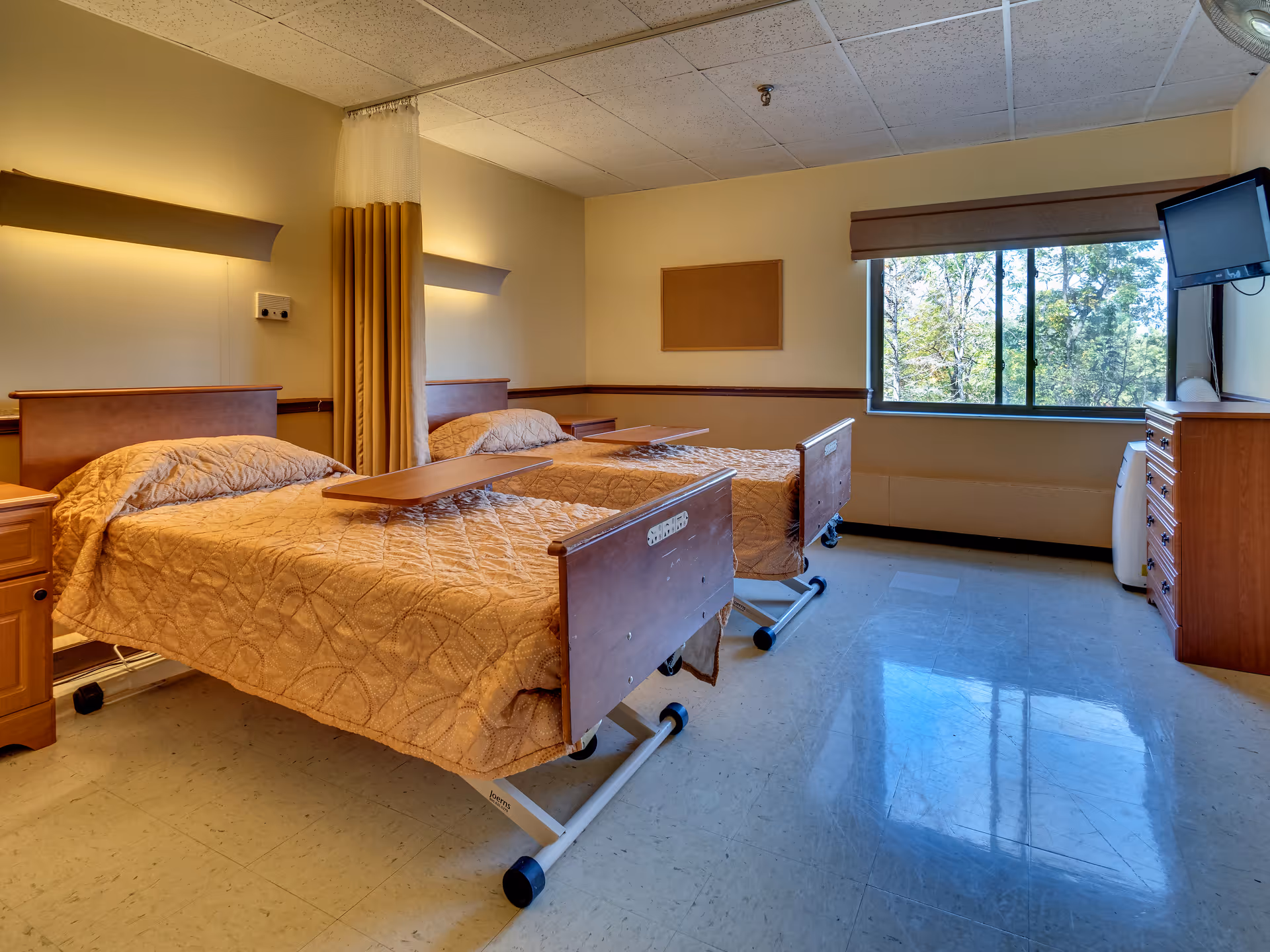 A shared senior care bedroom with two hospital-style beds, bedside tables, a window, and a wall-mounted TV.