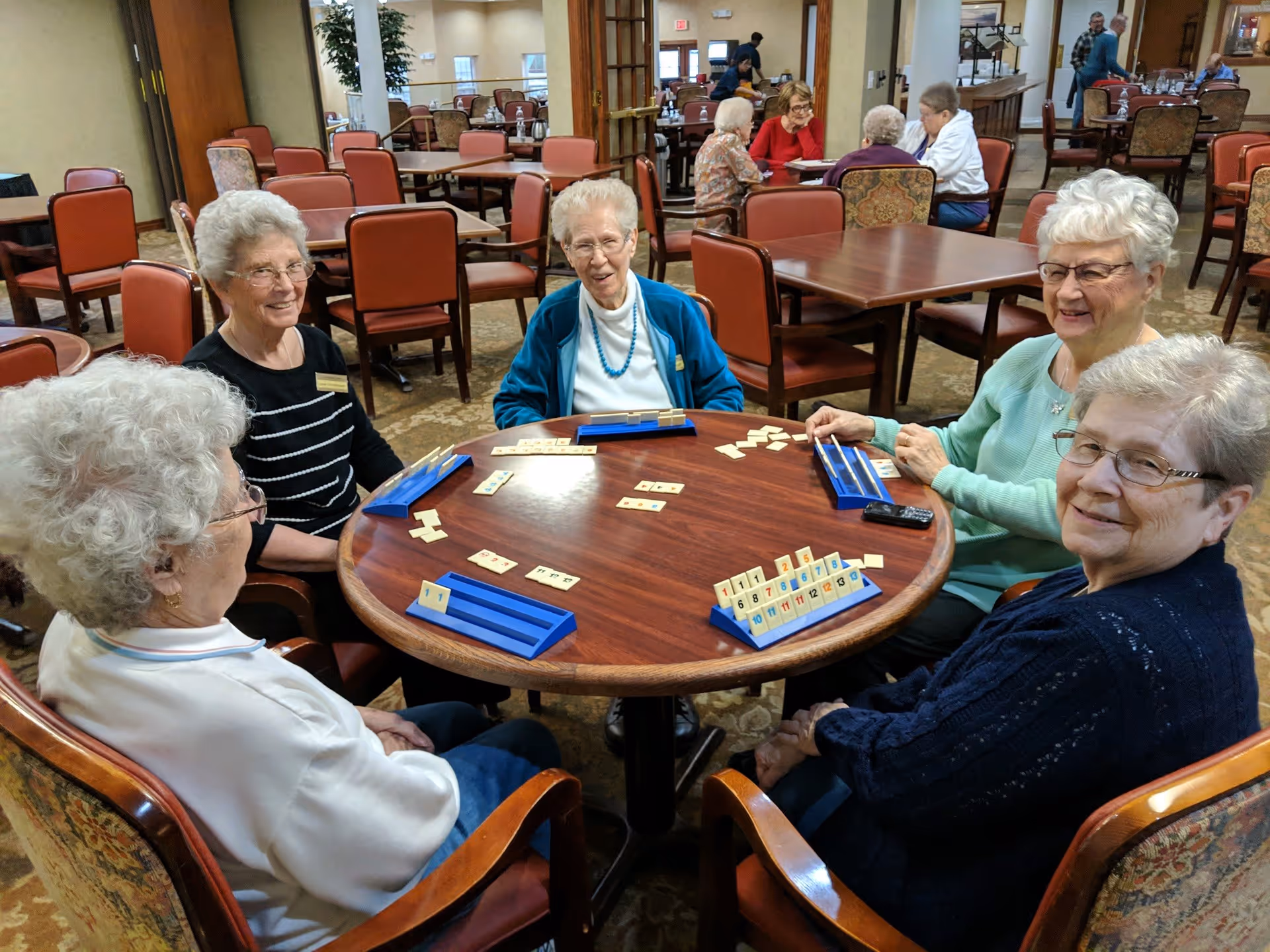 Five elderly women sitting around a round wooden table playing a tile-based game in a spacious room with multiple tables and chairs. The room has carpeted floors and warm lighting, with other people visible in the background also seated and engaged in activities.