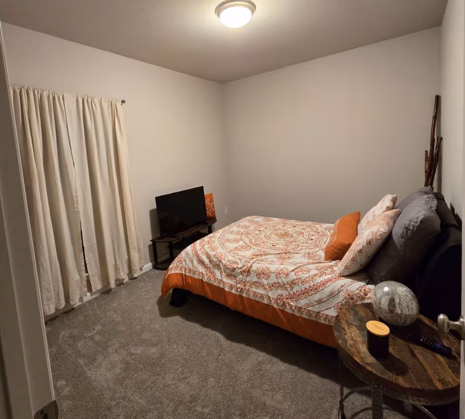 A small bedroom with a bed covered in a patterned orange and white bedspread, two pillows, and a gray headboard. There is a small wooden side table with a decorative stone, a candle, and a remote control. A flat-screen TV sits on a small stand against the wall. Beige curtains cover the window, and the room has beige carpet and plain light-colored walls with a ceiling light fixture.