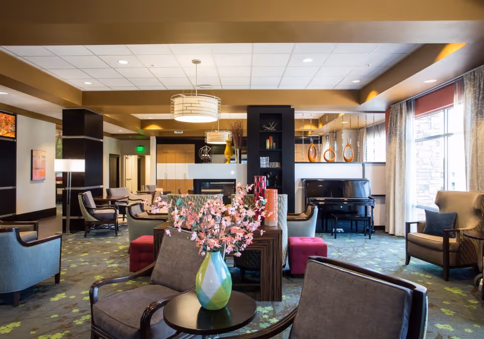 Bright, comfortable senior living lounge with multiple seating areas, a piano, and a central table holding a vase of pink flowers.