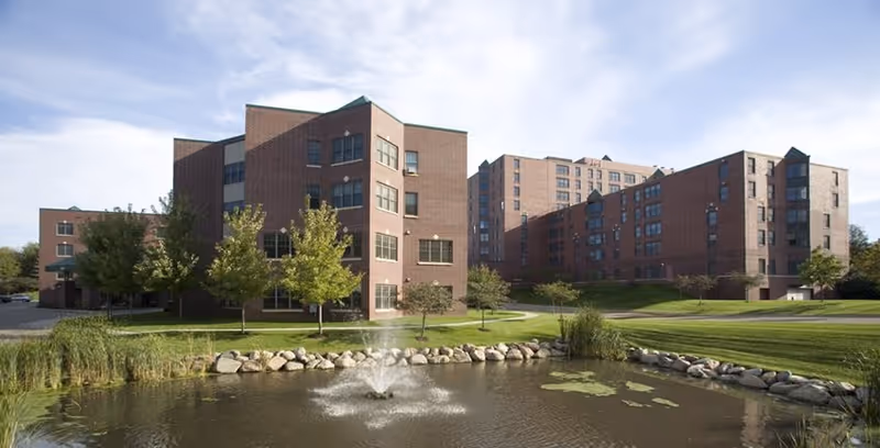 Exterior view of a large brick senior living facility with multiple connected buildings, surrounded by green lawns, trees, and a pond with a water fountain in the foreground under a partly cloudy sky.