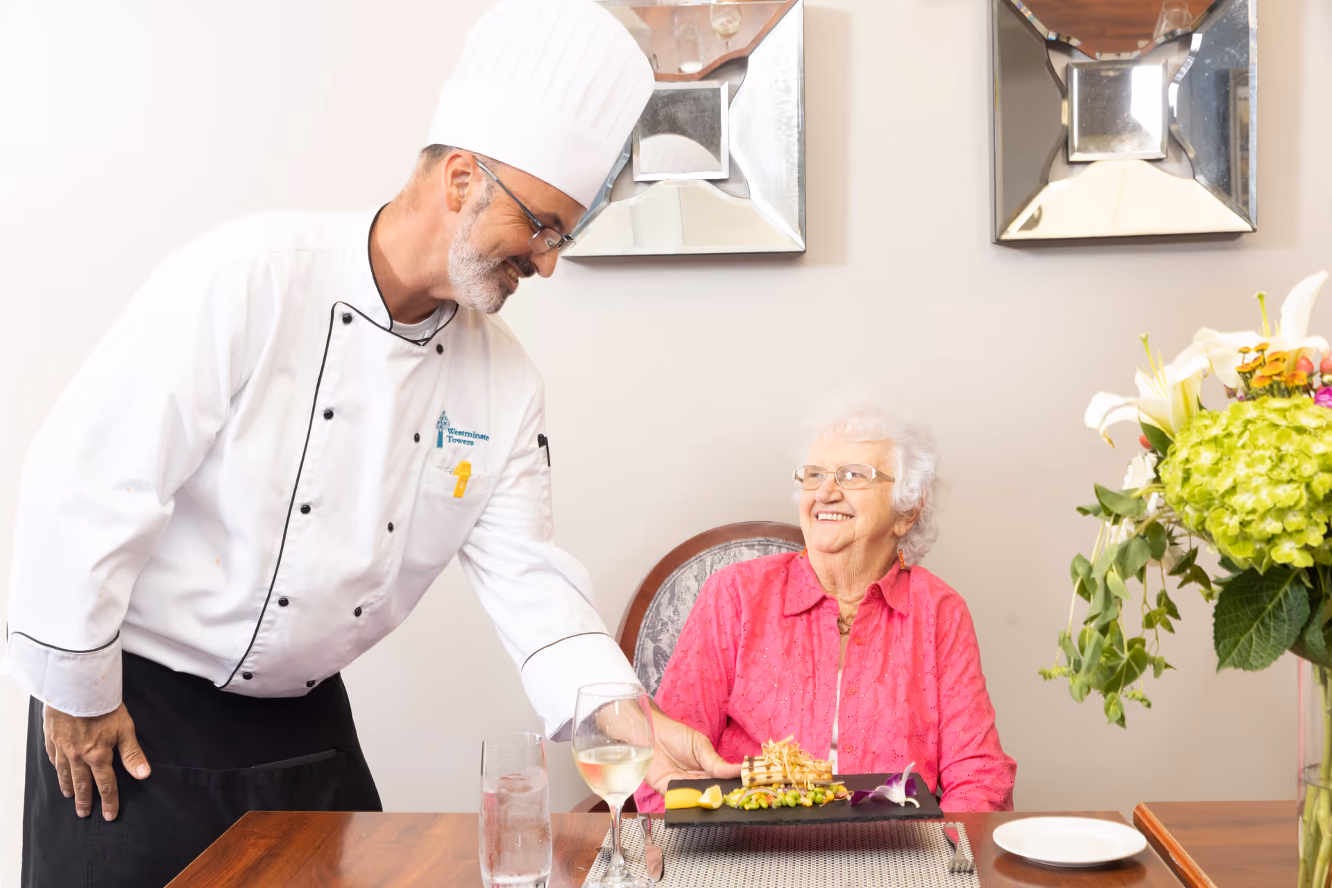 A chef in a white uniform serves a plated meal to a smiling elderly woman seated at a dining table.