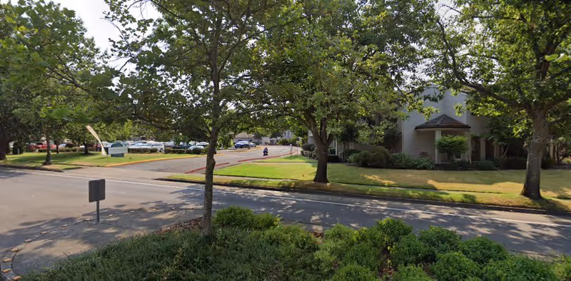 View of a street within a retirement community featuring green trees, bushes, and a grassy area. There is a building partially visible on the right side and several parked cars in the background. The scene is well-lit with sunlight filtering through the trees.