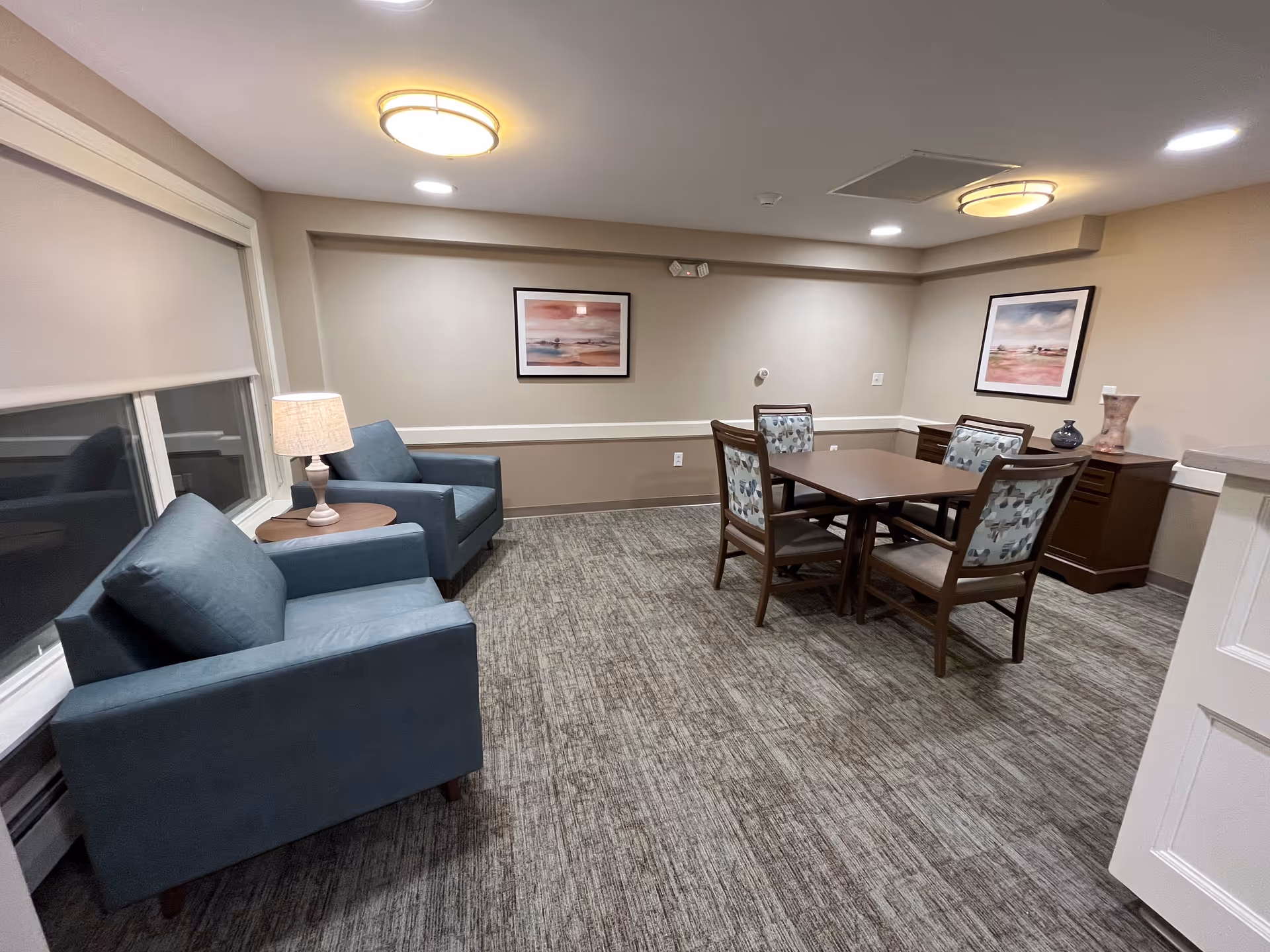 A furnished common room with blue armchairs, a table with four chairs, framed artwork, and soft ceiling lights.