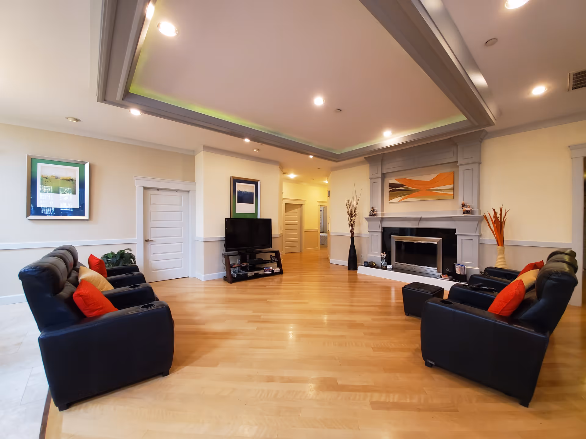 A spacious living room with light wood flooring, two black leather sofas facing each other with red and beige cushions, a modern fireplace with abstract artwork above it, a flat-screen TV on a stand, and decorative vases with tall dried plants. The ceiling has recessed lighting and a tray design with subtle green accent lighting.