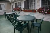 Outdoor patio area with green plastic tables and chairs arranged on a concrete floor. The patio is adjacent to a building with windows adorned with flower boxes containing pink and red flowers.