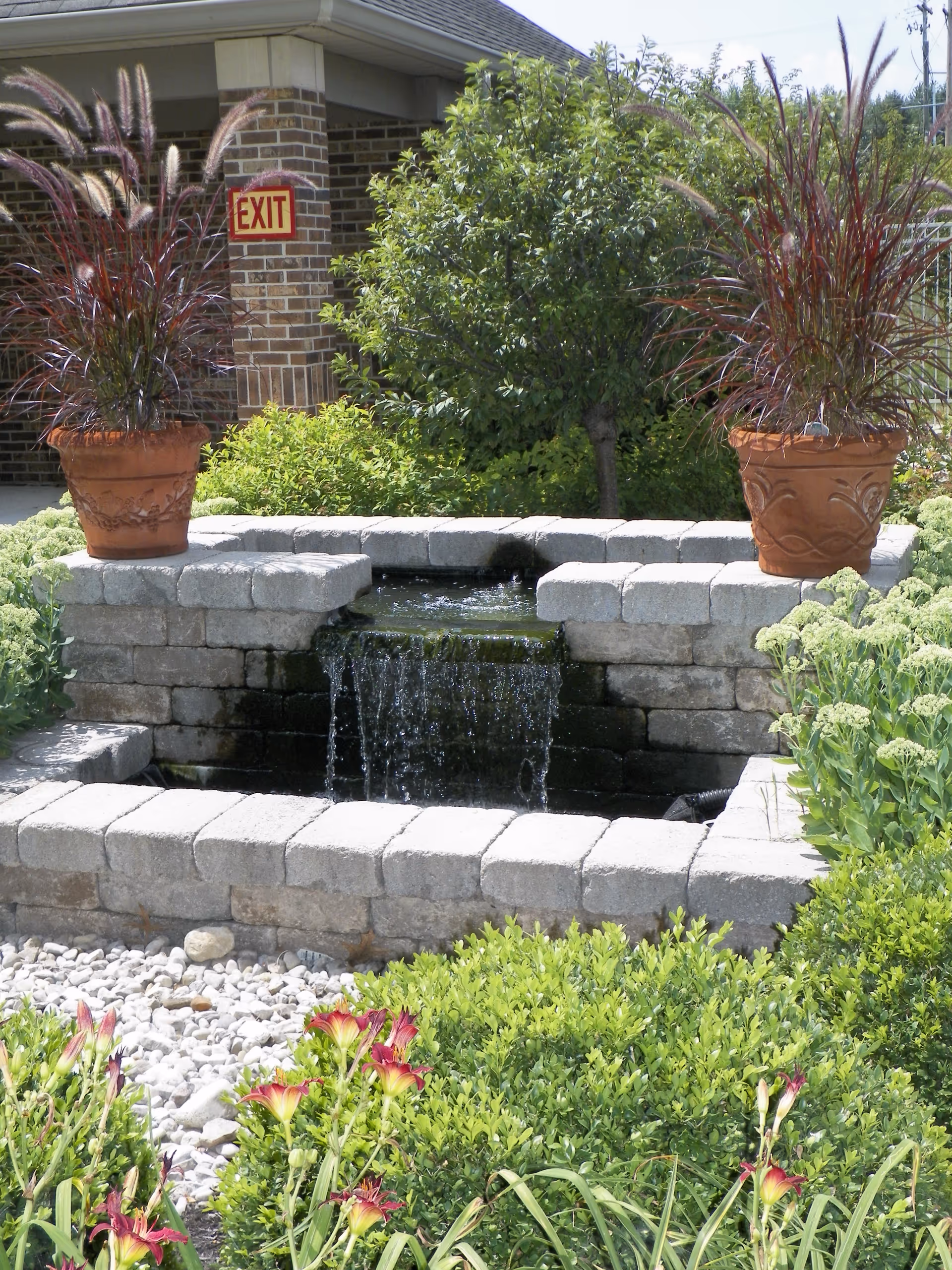 Outdoor garden area with a small stone water feature that has water cascading down a short ledge. Two large terracotta pots with tall ornamental grasses are placed on either side of the water feature. Surrounding the water feature are green shrubs, flowering plants, and a bed of white decorative stones. In the background, there is a brick building with an EXIT sign visible on a column.