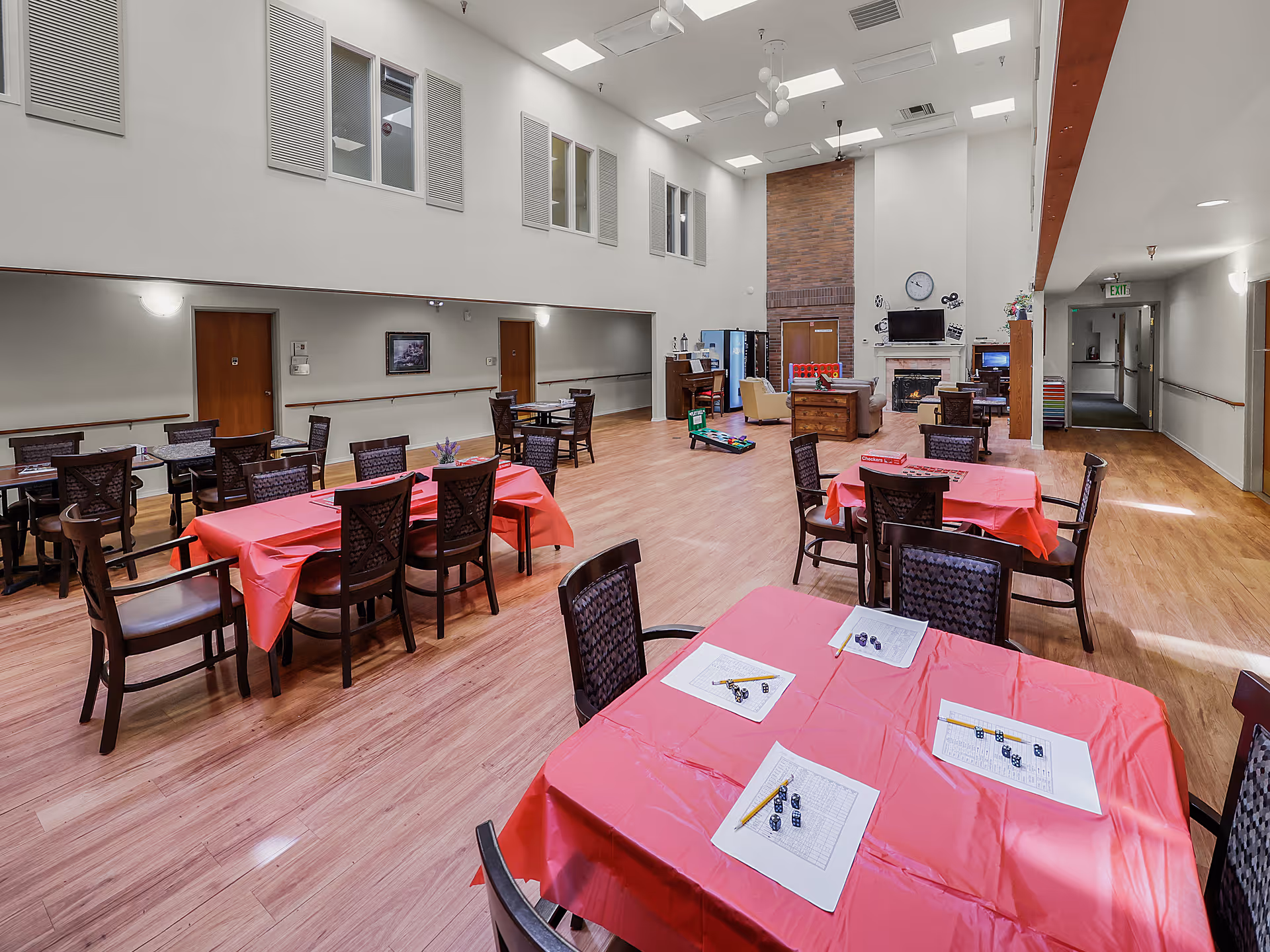 A spacious common dining/activity room with red-tableclothed tables and chairs, high ceilings with skylights, and a fireplace seating area.