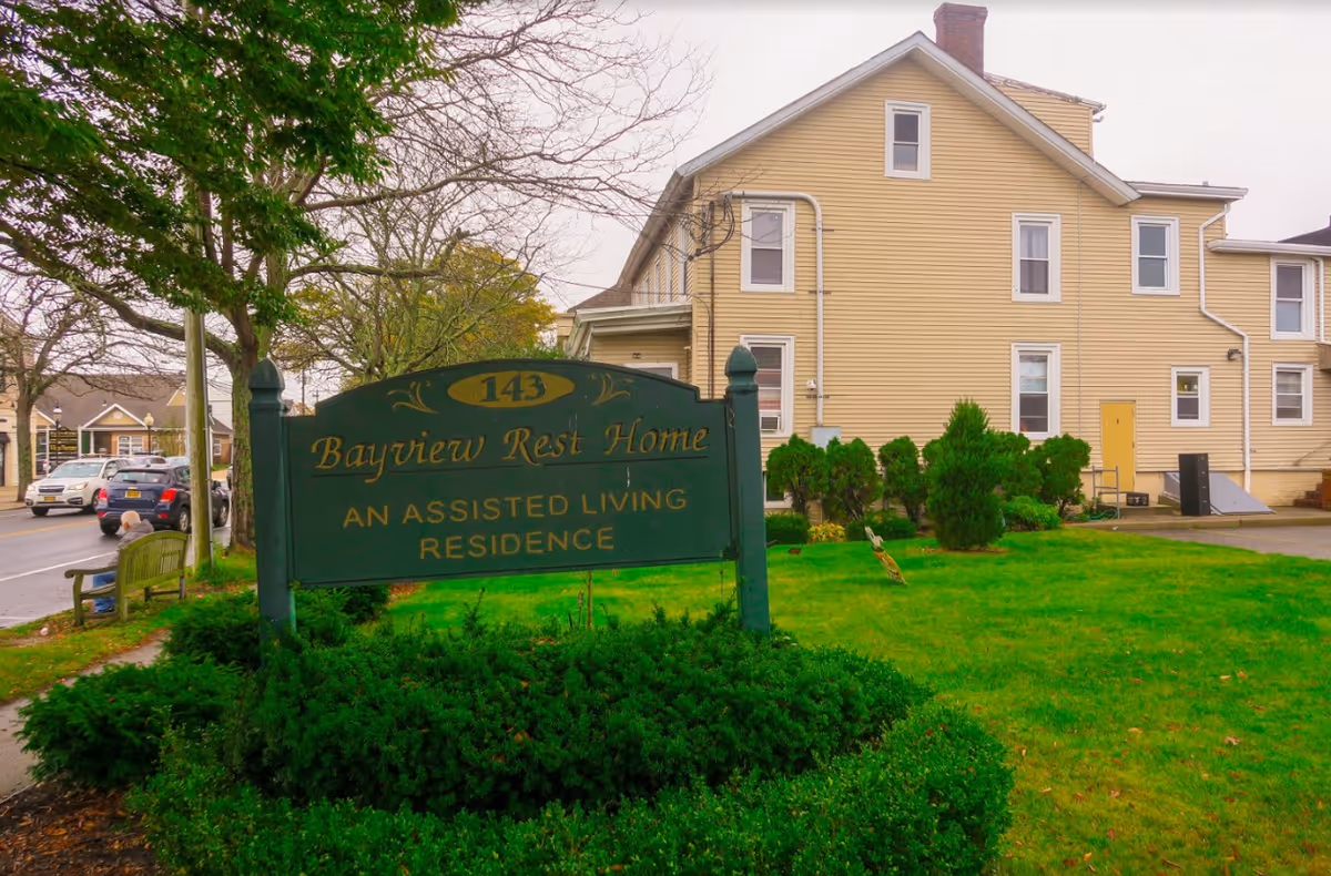 Exterior view of Bayview Rest Home, an assisted living residence, showing a yellow multi-story building with several windows and a green sign in the foreground displaying the facility's name and address number 143. There is a well-maintained lawn and some bushes around the sign, with a street and parked cars visible to the left.