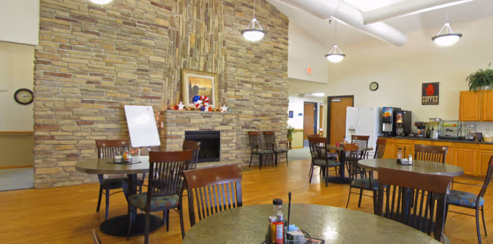Communal dining area with round tables and chairs, a large stone fireplace wall, and a coffee/refreshment station.