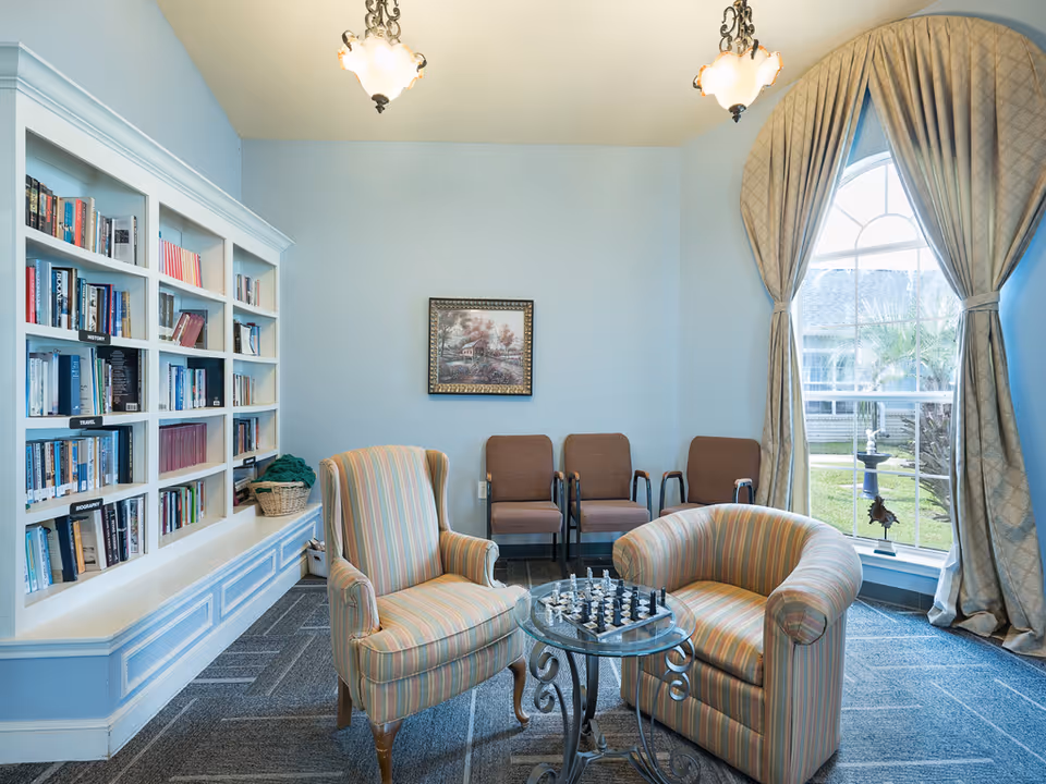 Cozy sitting area with striped armchairs around a glass chess table, bookshelves, additional chairs, and a large window with curtains.