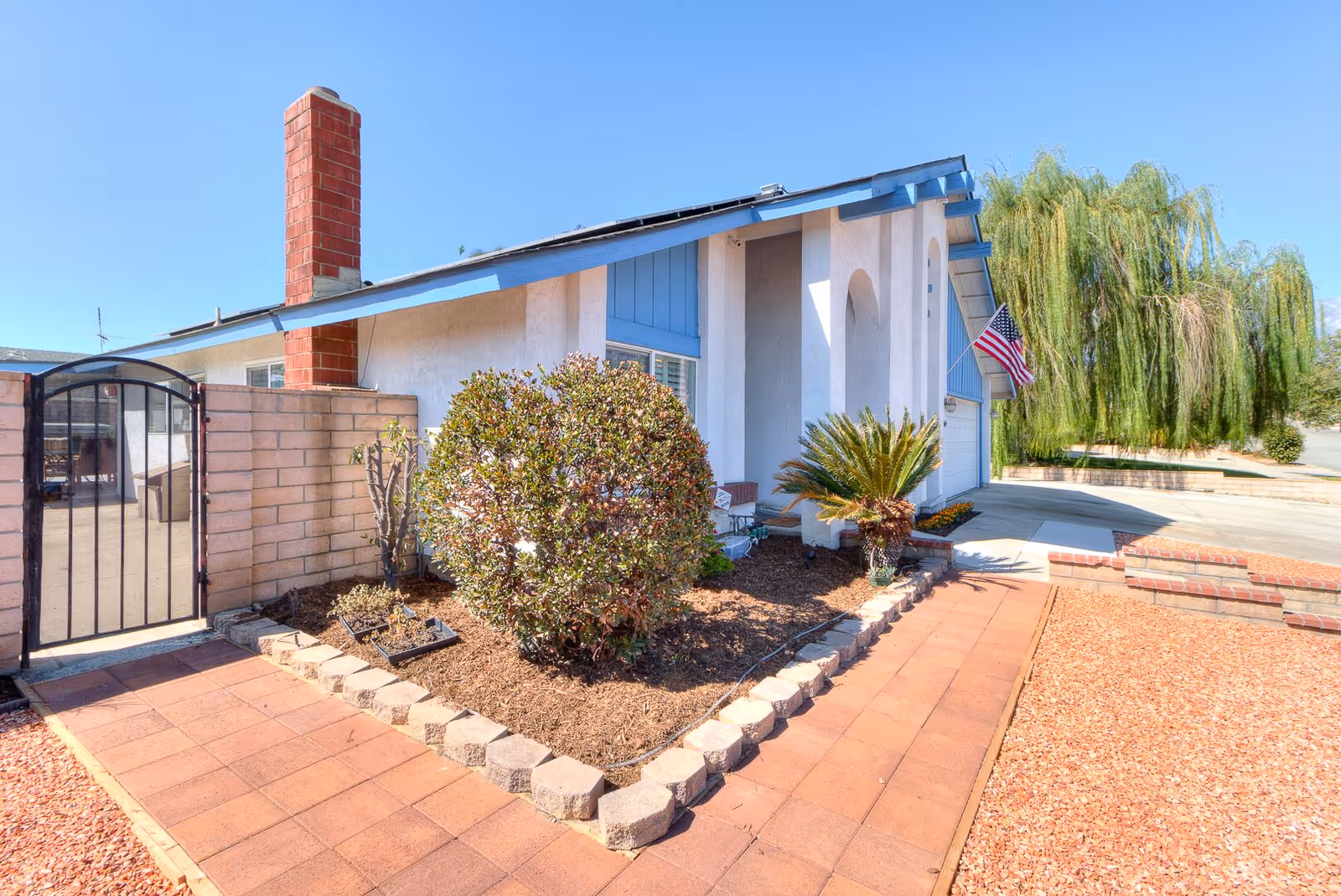 Front exterior view of a single-story house with a brick chimney, a small garden with shrubs and a palm plant, a paved walkway, and an American flag mounted near the entrance under a clear blue sky.