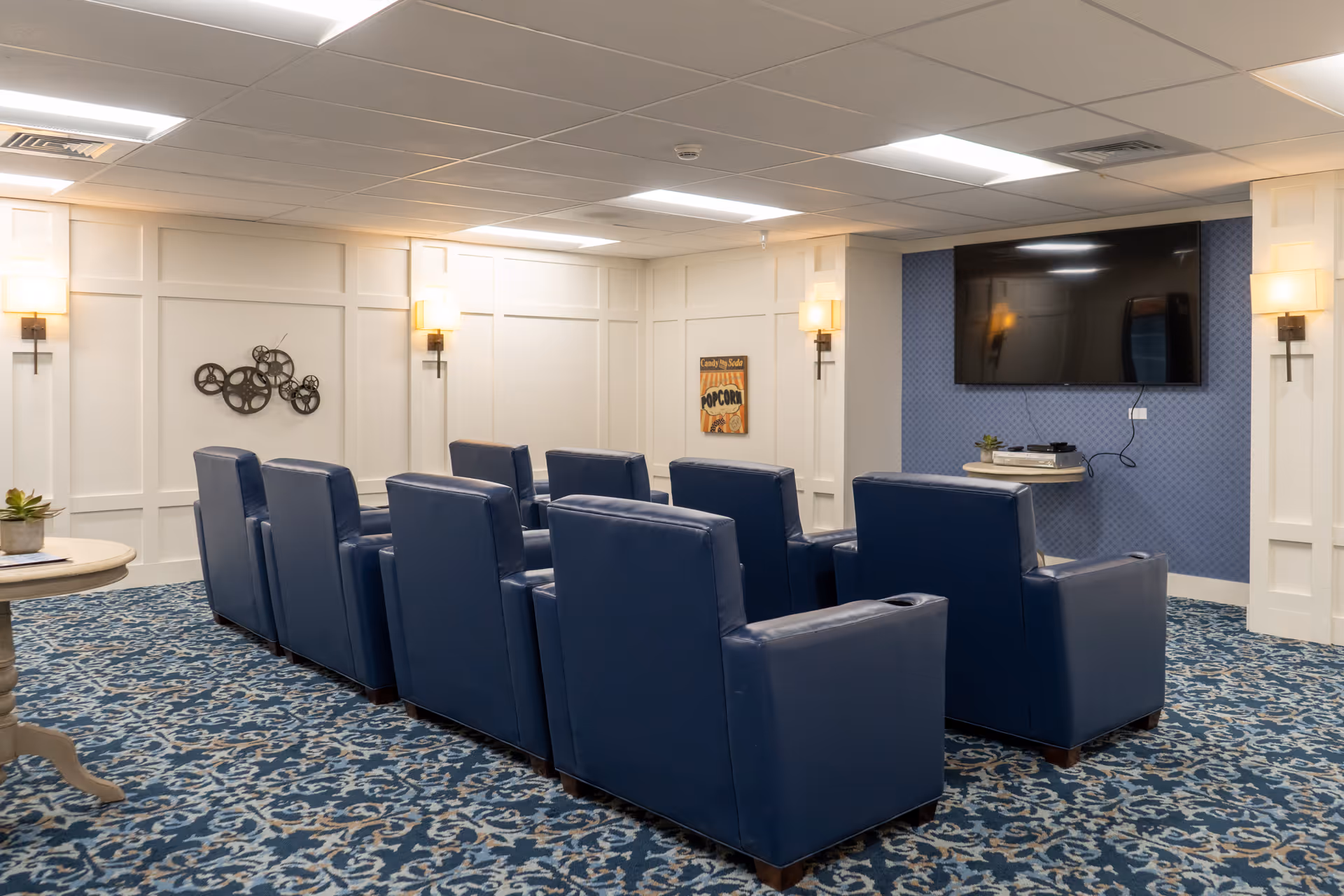 A cozy media room with two rows of blue leather recliner chairs facing a large flat-screen TV mounted on a blue accent wall. The room has patterned blue carpet, white paneled walls with wall sconces, and decorative wall art including a metal gear sculpture and a vintage popcorn sign.