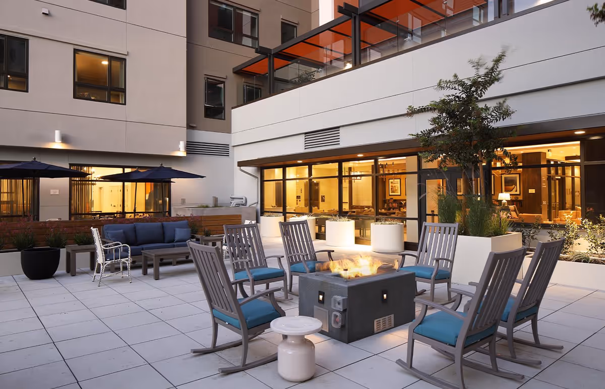 Outdoor patio area at Watermark at San Jose featuring a fire pit surrounded by eight wooden rocking chairs with blue cushions. There is a blue cushioned sofa and a white chair under two large black umbrellas. The patio is tiled and adjacent to a building with large windows showing a warmly lit interior.