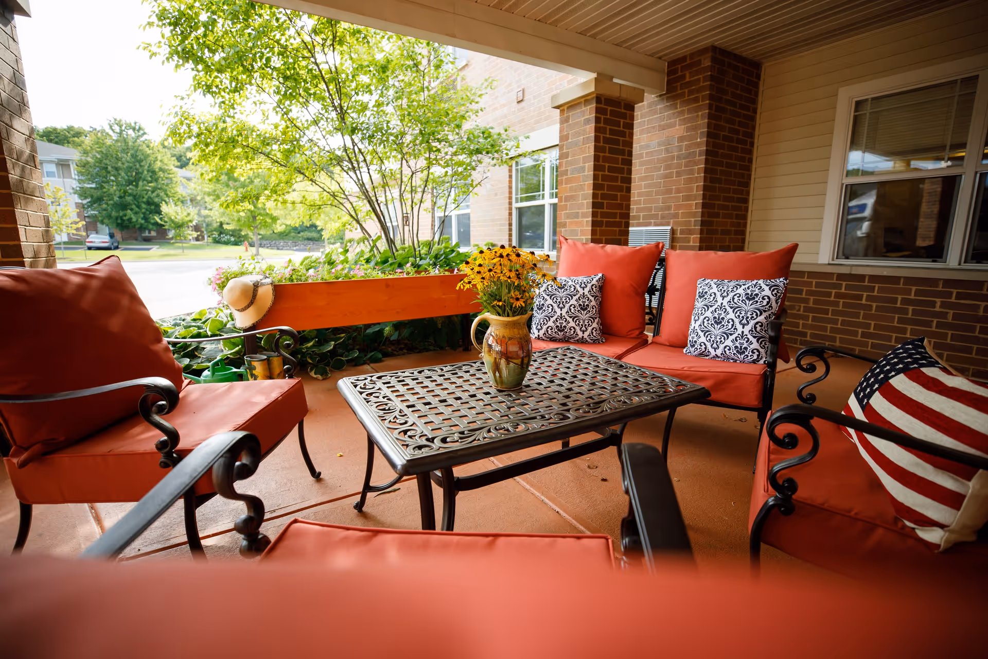 Covered outdoor patio area with red cushioned chairs and a metal table with a vase of yellow flowers. There are decorative pillows on the chairs, including one with an American flag design. A straw hat hangs on the wooden railing, and greenery is visible outside.