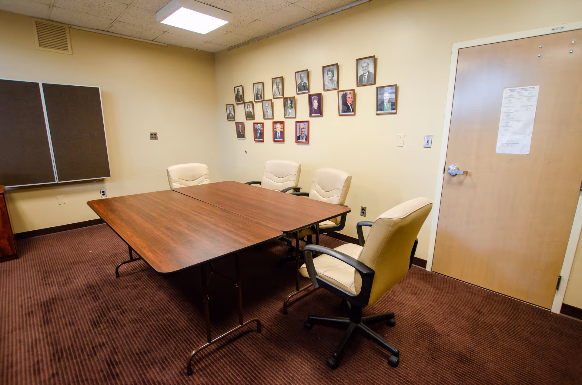 Small conference room with a rectangular wooden table, four beige swivel chairs, and framed portraits on the wall.
